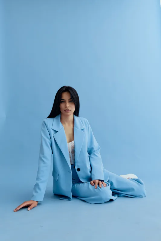 Young woman sitting on blue floor wearing light blue suit and white lace top, with long dark hair and neutral expression on soft blue background, minimal studio setting.