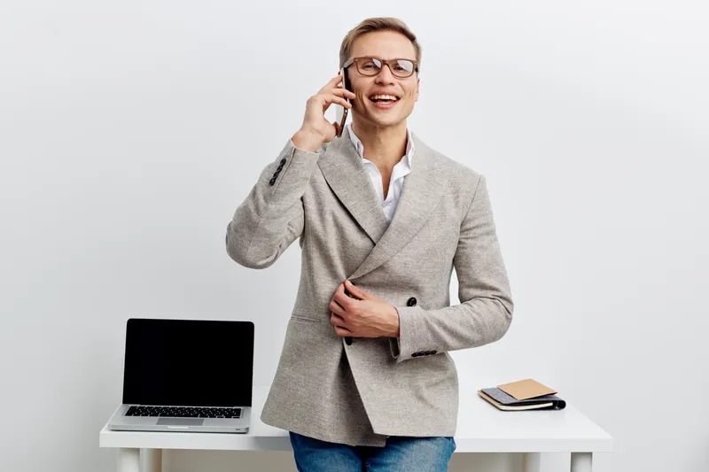 Smiling man in smart casual outfit talking on mobile phone while standing at office desk with laptop, notebook, and pen, isolated on plain white background. Business lifestyle concept.