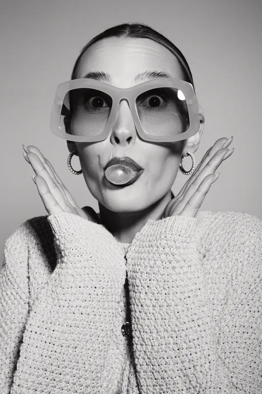 Playful Caucasian woman with pale skin wearing large glasses and knitted sweater makes funny face with puckered lips and hands near cheeks. Concept of humor, fashion, and casual lifestyle.