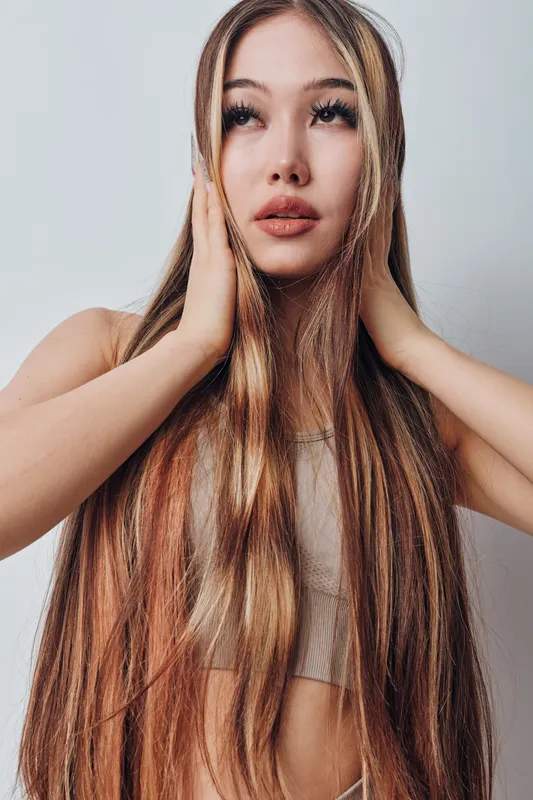Young woman with long straight hair and natural makeup poses with hands on head against white background. Beauty concept with healthy hair and thoughtful expression in studio light.