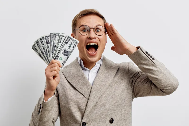 Eager excited middle-aged man with glasses, holding fan of dollar banknotes, wearing beige suit, smiling broadly, expressing happiness and wealth, isolated on plain light gray background, studio portr