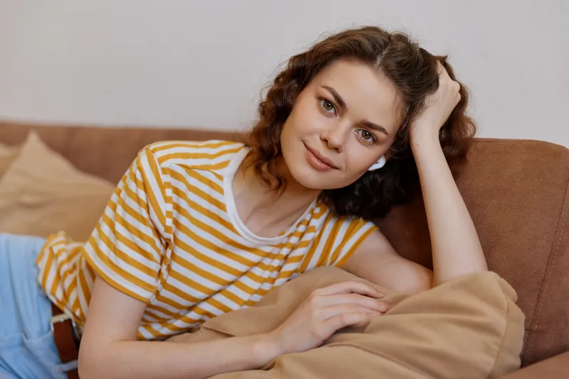 Relaxed woman with wavy hair lounging on a sofa wearing a striped t shirt, home setting. Lifestyle of comfort, casual mood, self care, and cozy ambiance with warm light.