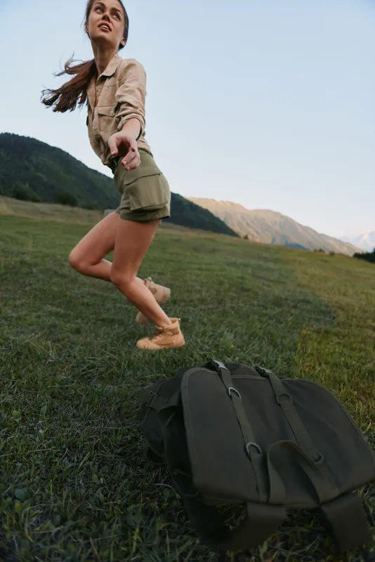 Young woman in shorts and jacket jumps joyfully on a grassy field with mountains in the background during an outdoor adventure.