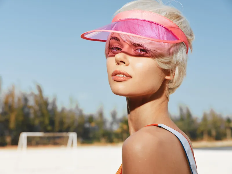 Bright portrait of a stylish woman wearing a pink translucent visor, sunlit skin, and a sporty summer vibe on an outdoor court under a clear blue sky.