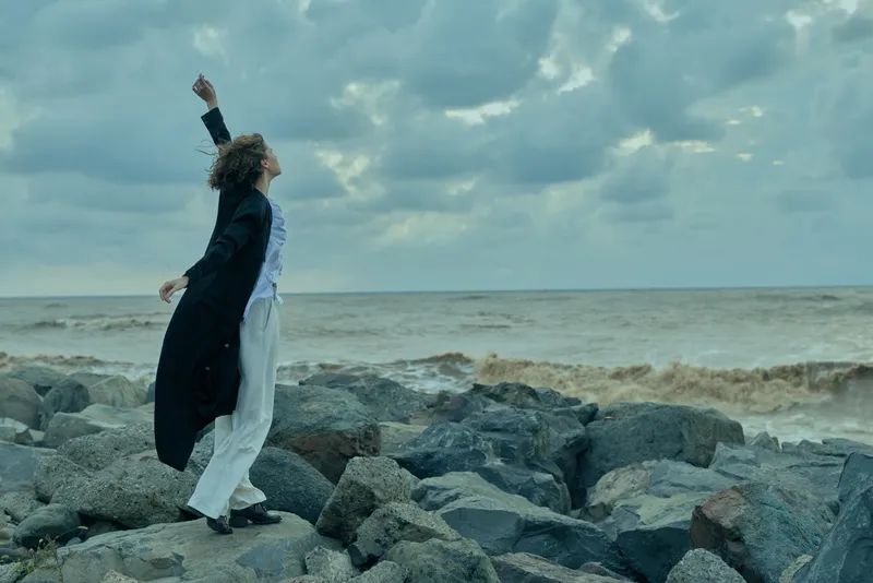 Woman in businesswear stands on a rocky coastline with arms raised in triumph, ocean breeze and dramatic sky conveying achievement, empowerment, and resilience during an outdoor moment.