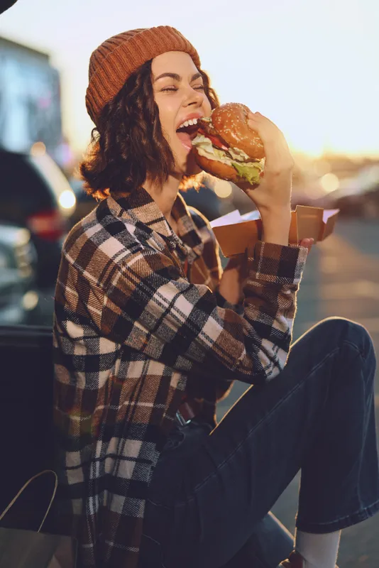 Woman eating burger outdoors at sunset, beanie and plaid jacket, smiling while taking a big bite. Authenticity, candid lifestyle and golden hour glow convey mindful living and emotional storytelling.