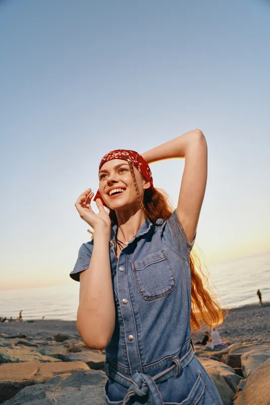 lifestyle woman wearing modern boho-western denim street style with a red bandana, smiling joyfully by the beach in warm film color tones at sunset