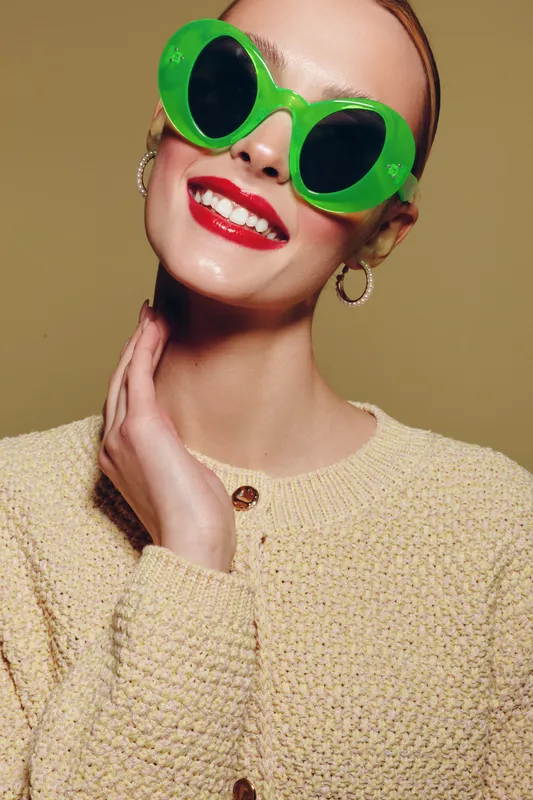 Smiling woman with bright red lipstick wears large green sunglasses and beige knitted sweater. Studio portrait on neutral background. Concept of fashion, style, and confident beauty.