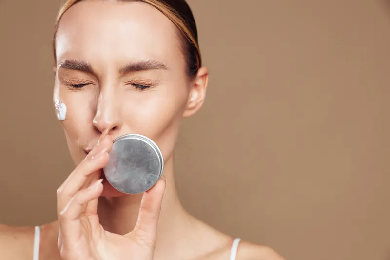 Young woman with closed eyes softly applies white facial cream to her cheek using a small applicator, captured in a tight close-up against a neutral beige background.