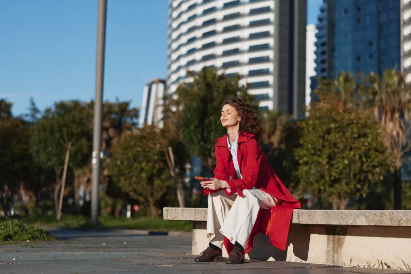 Cheerful woman outdoors, sitting on a bench in a red coat, enjoying sunlight, urban park setting, relaxed pose, natural colors, carefree mood