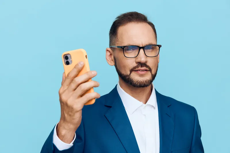 A professional man in a blue suit stands on a solid colored background, taking a confident selfie. He projects focus, poise, and ambition, embodying modern business leadership in a clean, minimal