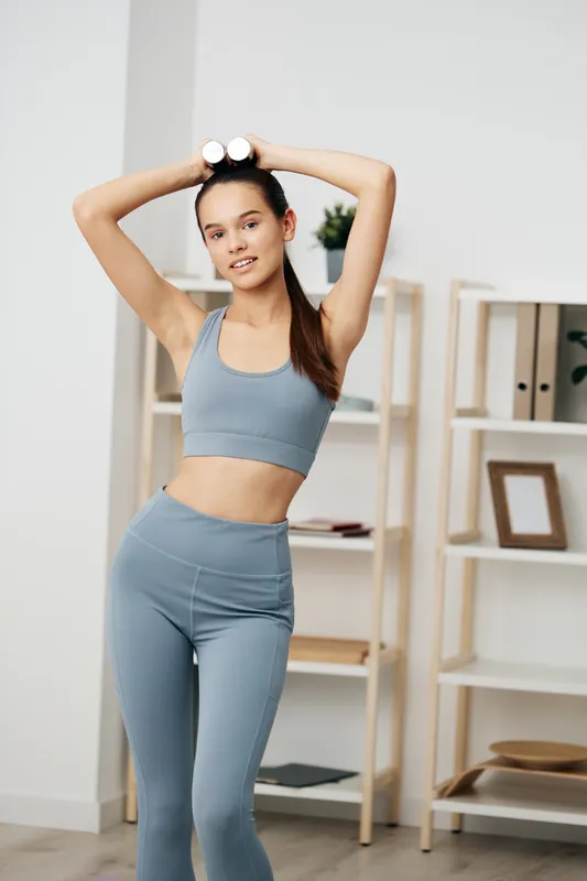 Fitness woman in blue athletic wear uses dumbbells overhead for a shoulder press at home gym, showcasing a focused workout and healthy lifestyle.