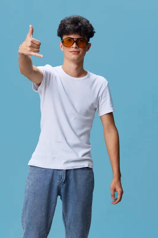 young man wearing sunglasses and casual white t-shirt shows shaka sign with hand, standing relaxed against solid blue background, friendly urban youth style expression