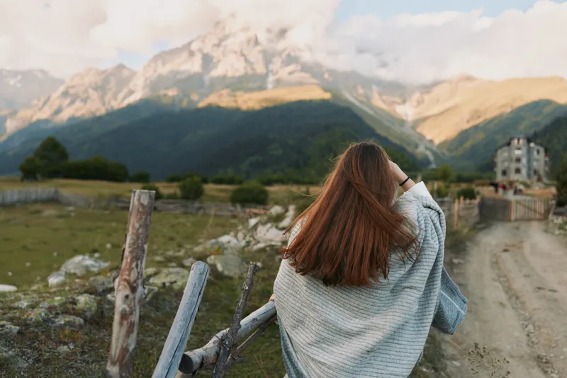 A woman stands with her back to the camera, overlooking rugged mountains from a wooden fence, capturing a serene, expansive landscape. Concept: solitude and nature.