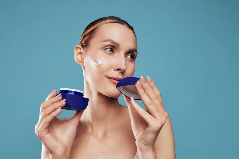 A young woman applies cream to her face from a blue jar in a bright studio, capturing a skincare routine concept focused on self care and beauty.