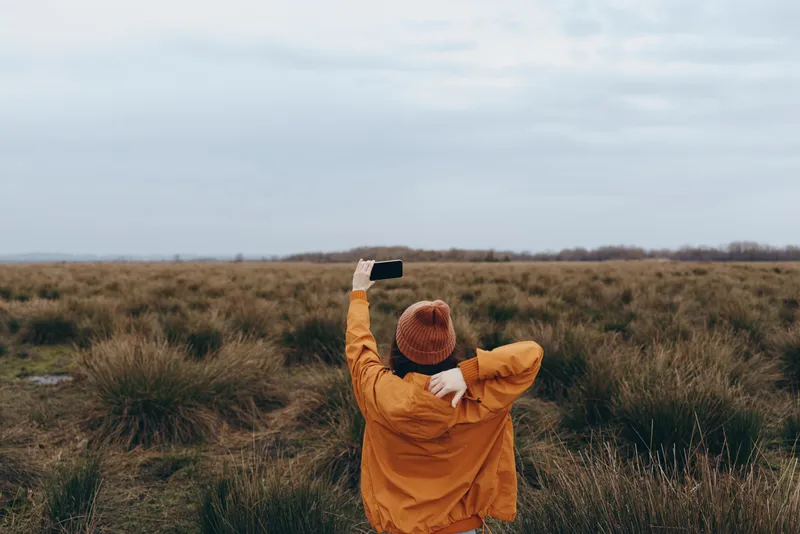 Woman lifestyle outdoors in a vast grass field, wearing orange jacket and hat, arms raised toward cloudy sky, enjoying quiet freedom, natural scenery and soft daylight today.