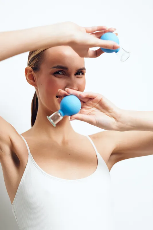 Young woman in white tank top holds blue facial suction cups near face, demonstrating skin care routine with clear skin and focused expression on white background.