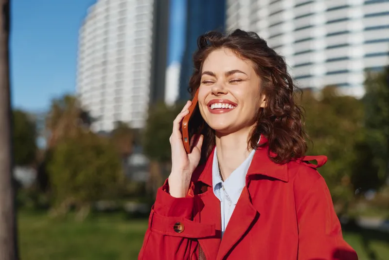 Happy woman in red coat talking on smartphone outdoors, smiling with closed eyes, enjoying sunny day in urban park with buildings in background.