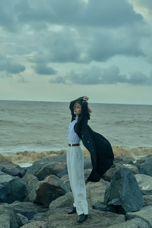 Woman on a rocky coastline, wind catching a long coat over white pants and striped shirt, as gray clouds sweep above the sea, creating a dramatic fashion moment for editorial stock and outdoor