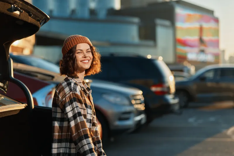 woman smile car parking beanie curlyhair candid golden glow in urban lot at golden hour, authenticity and candid lifestyle portrait with mindful living mood and emotional storytelling