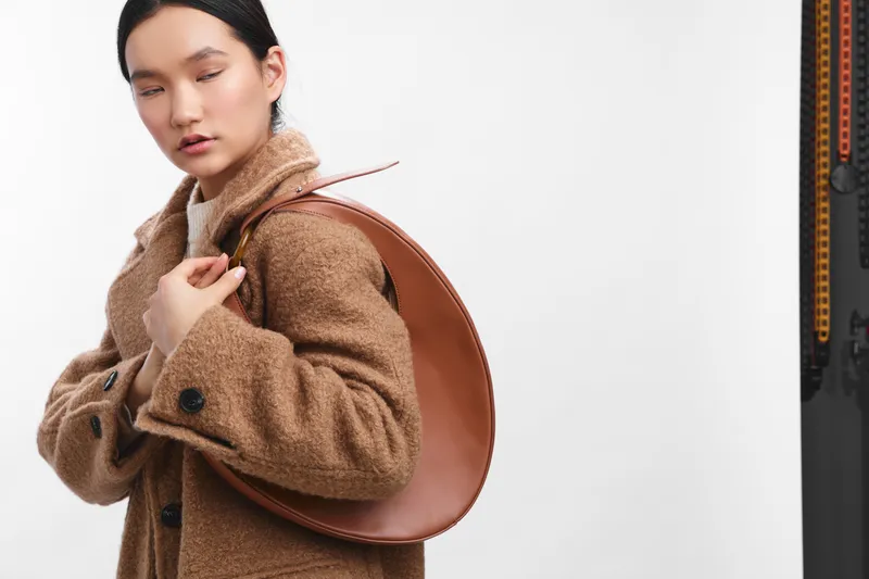 Asian woman wearing a brown coat with a round leather bag slung over her back, standing in a studio with a neutral backdrop, calm expression, poised and stylish.
