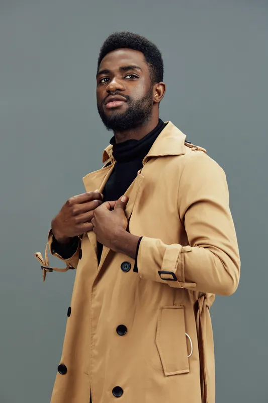 Stylish black male model in a beige trench coat against a minimalist grey background, showcasing confidence and elegance
