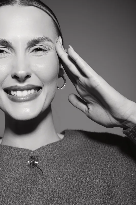 A smiling Caucasian woman with fair skin touches her face and smiles gently, wearing hoop earrings and textured sweater. Concept of beauty, happiness, and skincare.