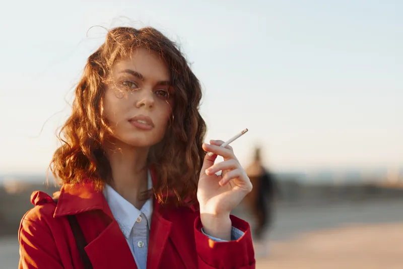 Young woman with curly hair wearing a red coat smokes a cigarette outdoors during golden hour with a calm expression and soft natural light on her face.