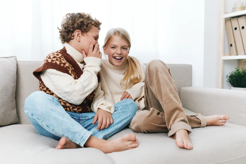 Two playful children share a secret on a cozy couch, smiling warmly as one whispers and the other giggles, creating a candid moment of friendship in a bright living room.