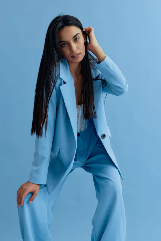 woman in blue suit posing confidently against a pastel blue background with long black hair and natural makeup, showcasing modern business fashion and style
