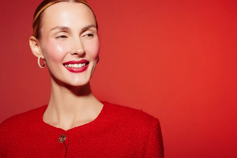 Smiling woman with shiny skin and red lipstick wears red sweater and silver hoop earrings. Portrait on red background with soft lighting. Concept of beauty, fashion, and confidence.