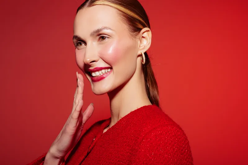 Smiling woman with shiny skin and red lipstick poses on red background wearing red outfit and gold earrings. Concept of beauty, fashion, makeup, and confident female portrait.
