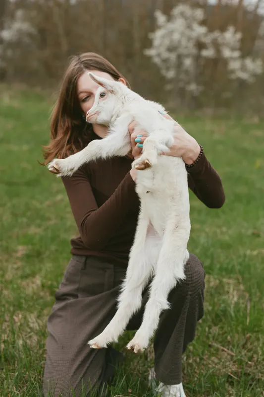 A young woman in brown clothes gently holds a white baby goat outdoors in a green field during spring, capturing a tender moment of connection and lifestyle.