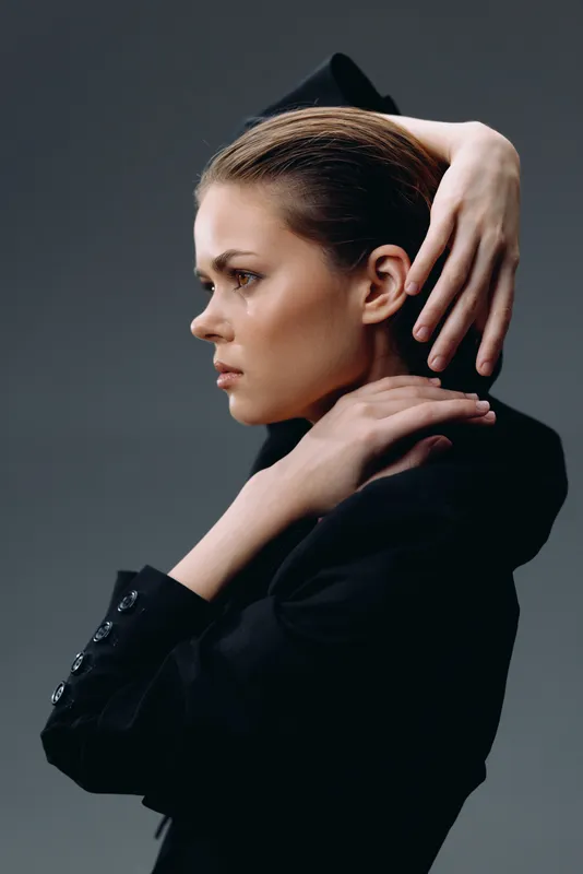 Profile of a thoughtful woman in black clothing with hands touching neck and head, minimalistic background, moody portrait mood.