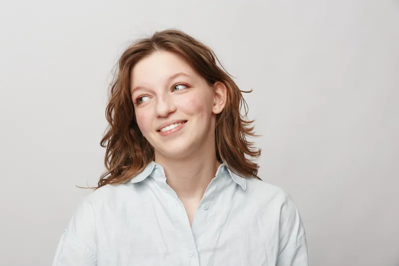 Smiling young woman with wavy brown hair wearing a white shirt looks sideways on a light background. Concept of happiness, casual style, and natural beauty portrait.
