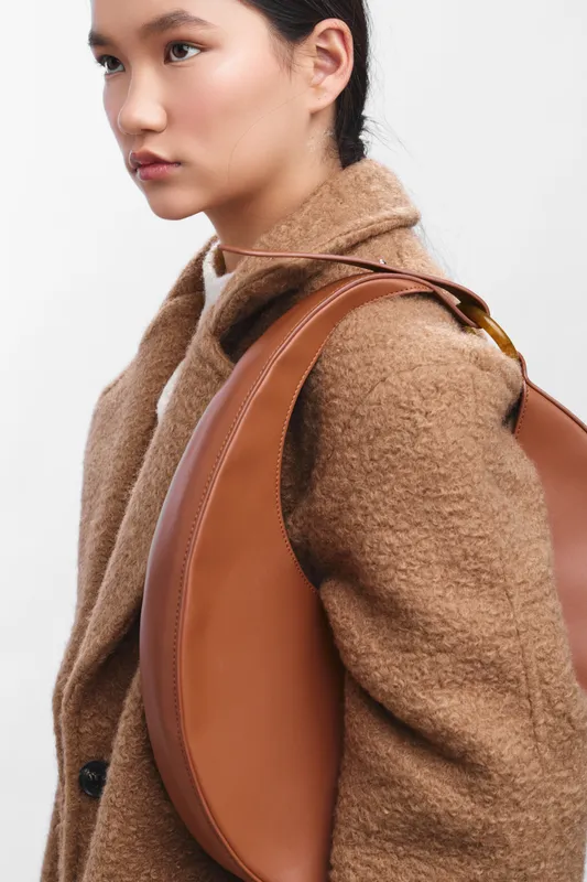 Asian woman wearing a warm brown coat and carrying a leather backpack stands in a clean, neutral setting, exuding confidence, style, and poise with a composed expression.