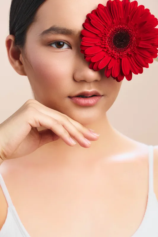 asian woman gazes softly in a studio portrait with a bright red flower tucked near her eye, creating an artistic and elegant focal point.