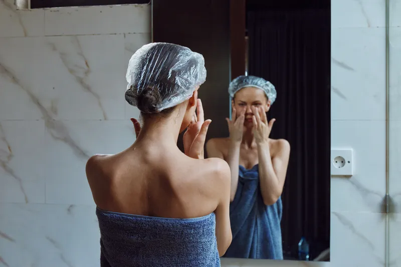 woman wearing shower cap applying skincare cream on face in bathroom mirror, healthy skincare routine, morning self-care, wellness concept