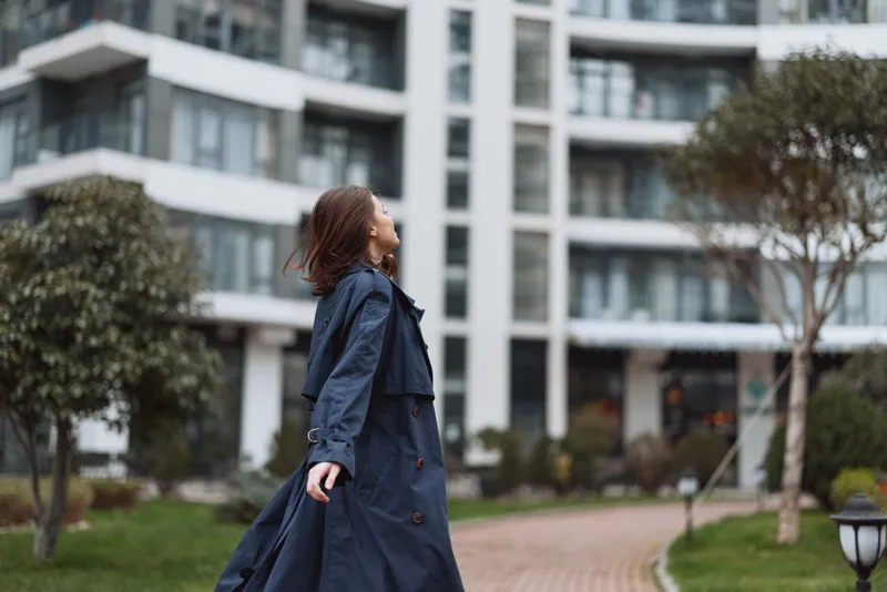Young woman in a long dark coat enjoys a walk in an urban park with modern buildings in the background during a calm day.