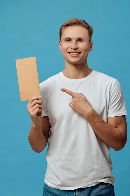 Smiling young man holding blank cardboard sign pointing at it, casual white t-shirt, blue background, studio portrait, happy expression, advertising concept. People lifestyle concept, cheerful, positi