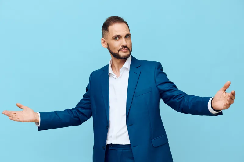 A professional man in a blue suit stands against an isolated colored blue background, arms open wide, conveying confidence, openness, and approachable leadership in a corporate setting.