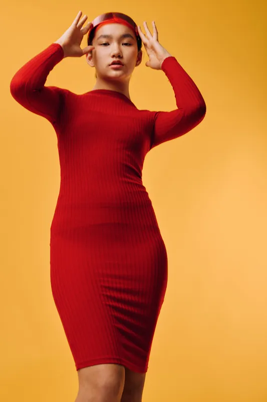 Young woman posing with hands near head wearing tight red dress and matching headband. Studio portrait with warm yellow background, fashion model, confident and stylish look for beauty concept.