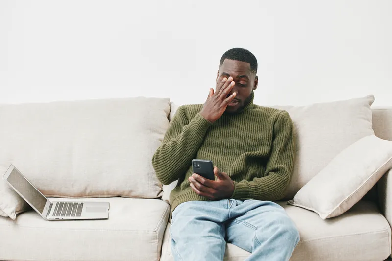 A stressed young Black man in a green sweater sitting on a sofa, using a smartphone while feeling overwhelmed by daily life challenges
