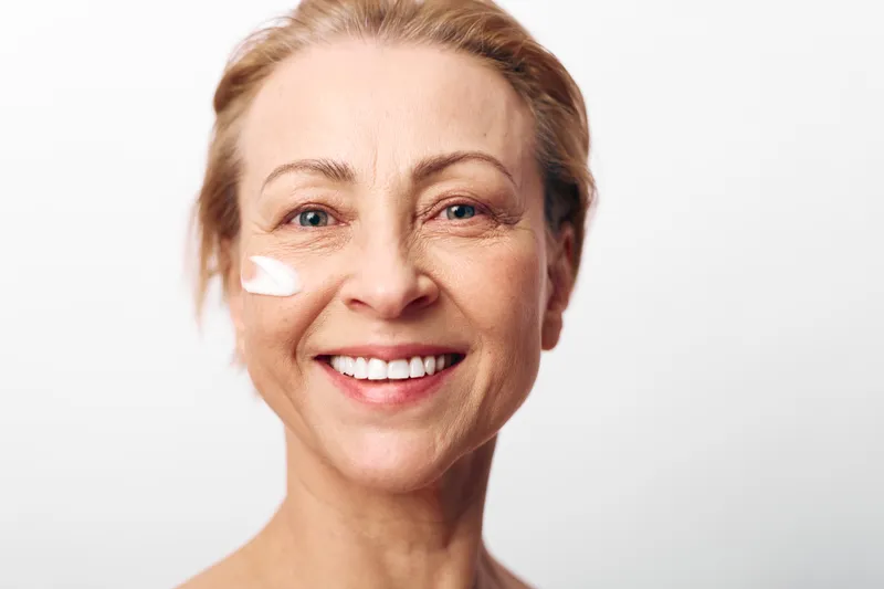 Smiling mature woman with natural skin applying white cream on her cheek, isolated on white background. Concept of skincare, beauty routine, anti-aging and self-care for middle aged women.