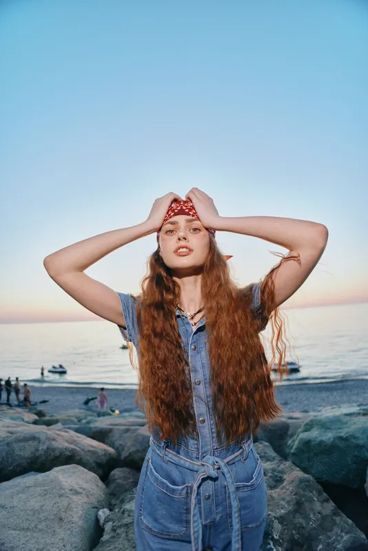 young woman with long hair wearing blue denim dress and red bandana standing on rocky beach at sunset with calm sea and clear sky in background