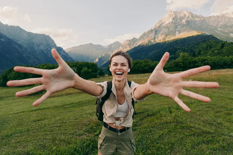 Joyful young woman with arms stretched toward the camera, smiling in a meadow under towering mountains, green grass, clear sky, and a sense of freedom.