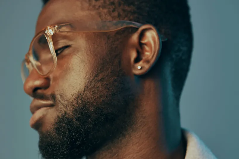 Thoughtful young Black man with glasses on a blue background, expressing calmness and introspection, ideal for concepts related to self reflection and mindfulness