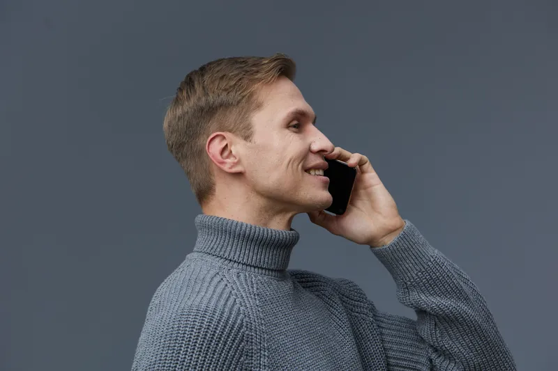 Smiling man talking on phone with a friendly expression, wearing a stylish gray turtleneck sweater, isolated on plain gray background, studio portrait with focus on facial features and communication c