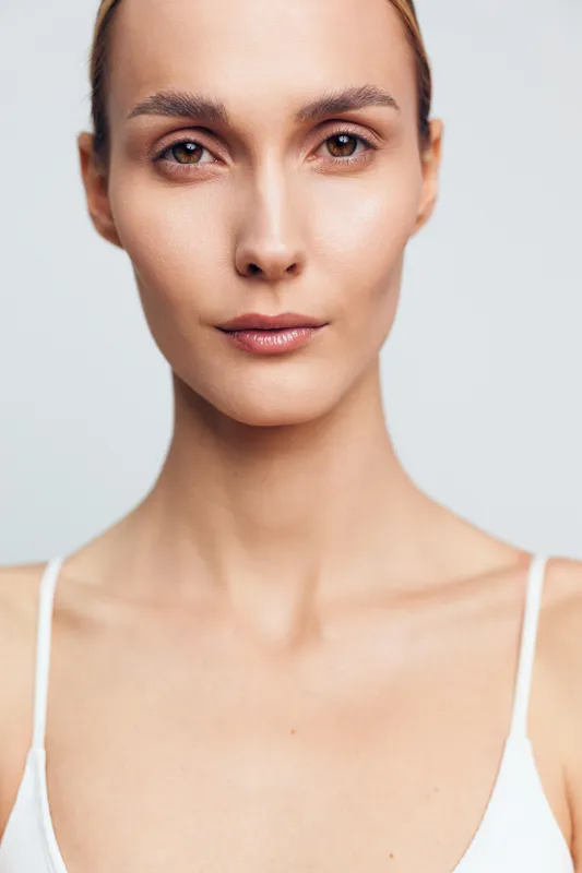 Closeup portrait of a woman with clear skin and natural makeup looking at camera, neutral expression, soft lighting, minimalistic background, wearing white top