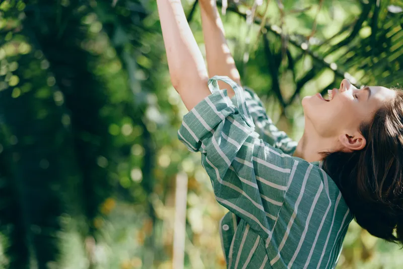 A woman on a swing outdoors, arms raised, wearing a striped shirt, sunlight filtering through green leaves, capturing a playful, carefree moment.
